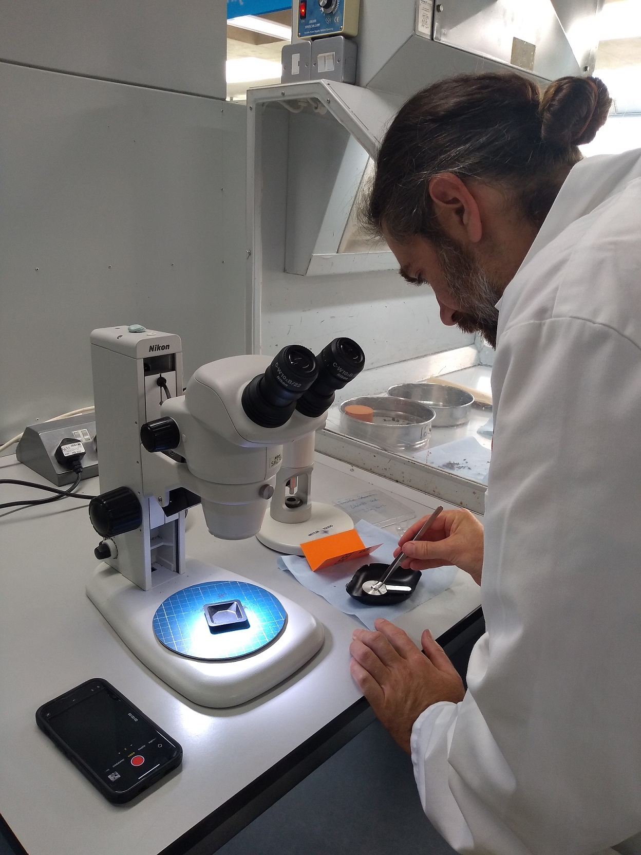 Dustin wearing a white lab coat, bent over a small black tray of seeds holding tweezers. Next to him on the bench is a microscope with a small black tray on ready for the next sample